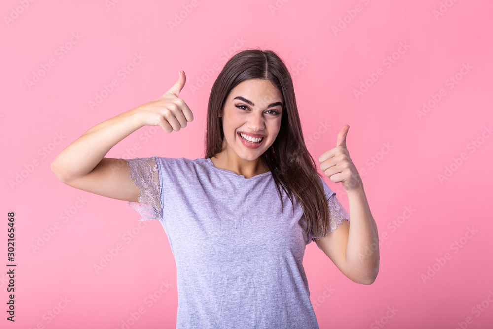 Smiling pretty young woman showing thumbs up isolated on the pink background. Smiling woman showing thumbs up and looking at the camera Smiling pretty young woman showing thumbs up isolated on the pink background. Smiling woman showing thumbs up and looking at the camera