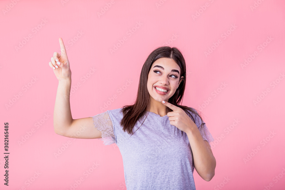 Portrait of brunette woman with long hair in basic t-shirt rejoicing and pointing finger at copyspace isolated over pink background