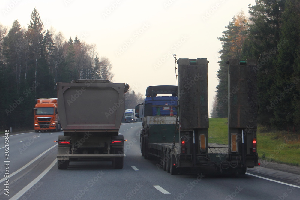 Heavy dump truck overtake of low-frame semi-trailer trawl truck on a two-lane asphalt highway forest road in the autumn evening, rear-side view – Logistics, transportation, trucking industry