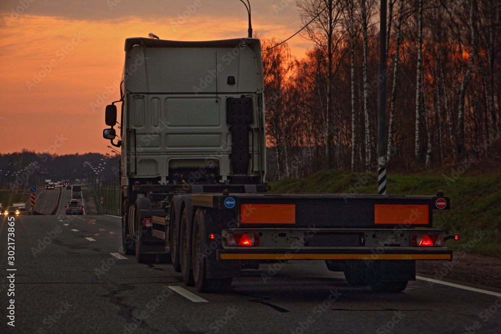 Empty heavy truck with low-frame semi-trailer trawl on a two-lane asphalt highway road in the beautiful autumn evening, rear-side view – Logistics, transportation, trucking industry