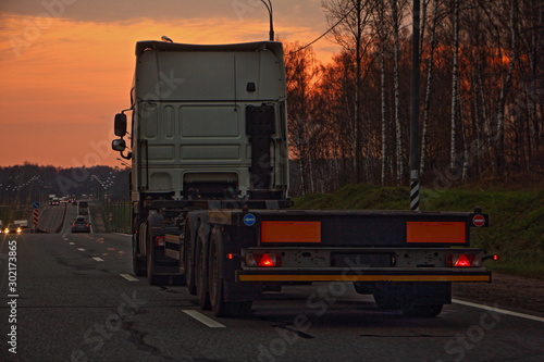 Obraz na płótnie Empty heavy truck with low-frame semi-trailer trawl on a two-lane asphalt highway road in the beautiful autumn evening, rear-side view – Logistics, transportation, trucking industry w salonie