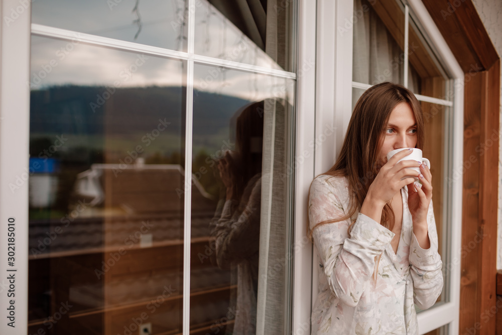 Portrait of a happy smiling young woman in stylish nightwear with a cup of coffee or tea meeting the day standing on the hotel terrace with a gorgeous view of the mountains. place for inscription Portrait of a happy smiling young woman in stylish nightwear with a cup of coffee or tea meeting the day standing on the hotel terrace with a gorgeous view of the mountains. place for inscription