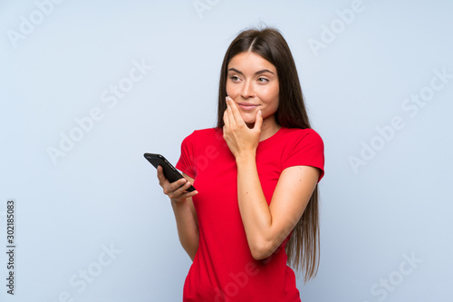 Brunette young woman with a mobile phone over isolated blue wall thinking an idea
