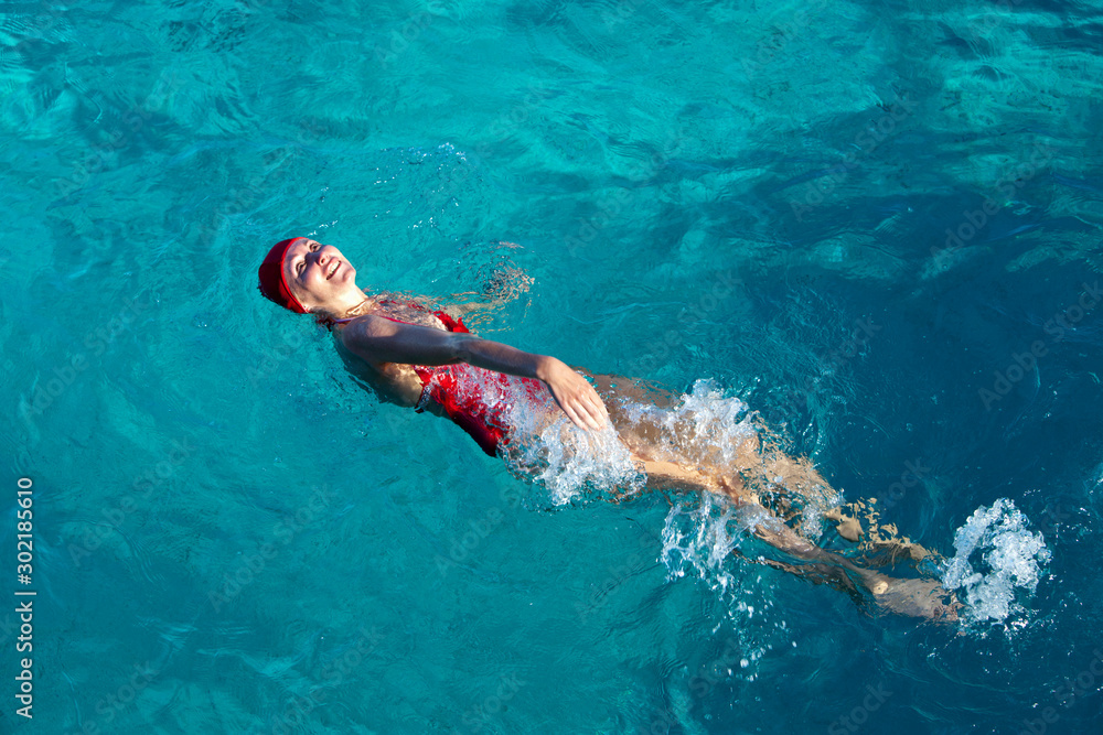 young woman in a red swimsuit and a red sports cap with a good slim figure swims on her back in a bright blue tropical sea and smiling
