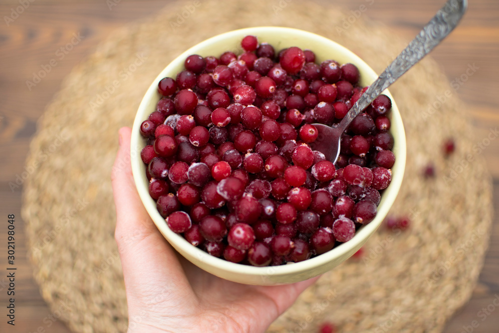 Female hand giving big bowl full of frozen cranberry with metal spoon inside at blurred wooden background with eco napkin, scattered berries, eco living Female hand giving big bowl full of frozen cranberry with metal spoon inside at blurred wooden background with eco napkin, scattered berries, eco living