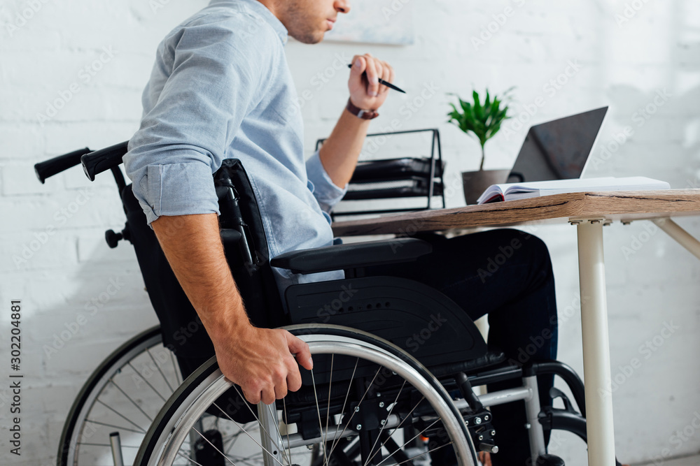 Cropped view of man sitting in wheelchair by notebook at workplace Cropped view of man sitting in wheelchair by notebook at workplace