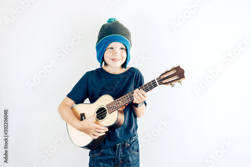 Portrait of a positive boy in casual clothes and hat playing ukulele. Copy space on white background.