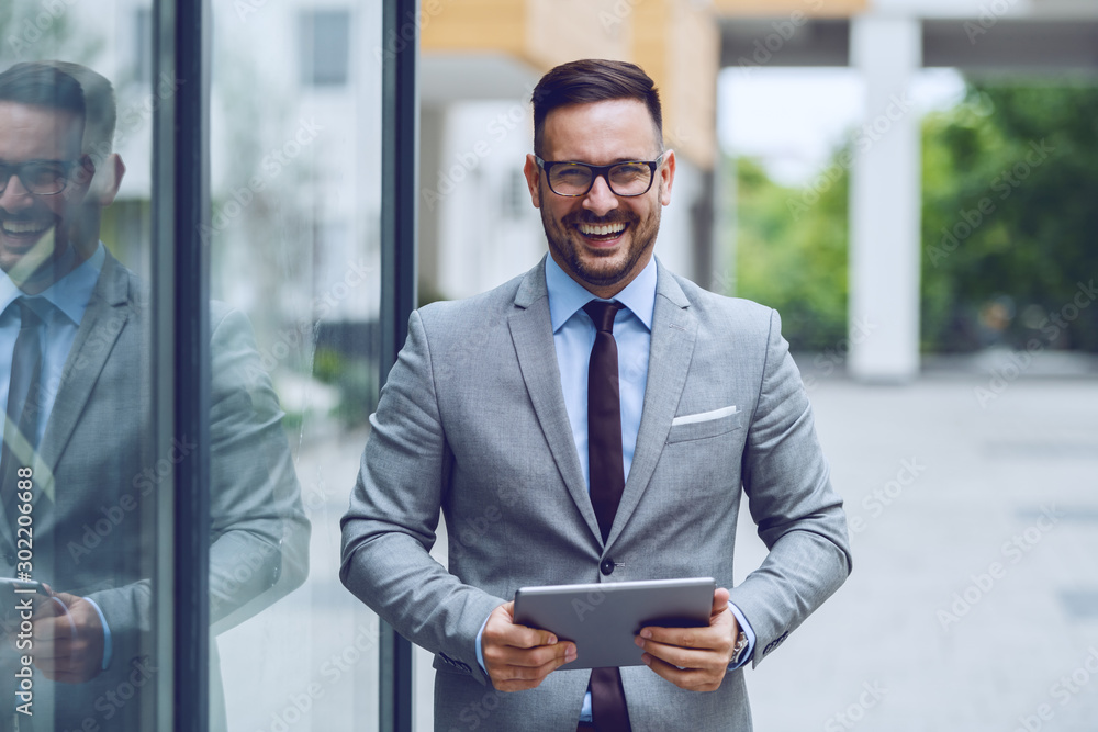 Handsome smiling caucasian businessman in gray suit and with eyeglasses holding tablet while standing outdoors and looking at camera. Handsome smiling caucasian businessman in gray suit and with eyeglasses holding tablet while standing outdoors and looking at camera.