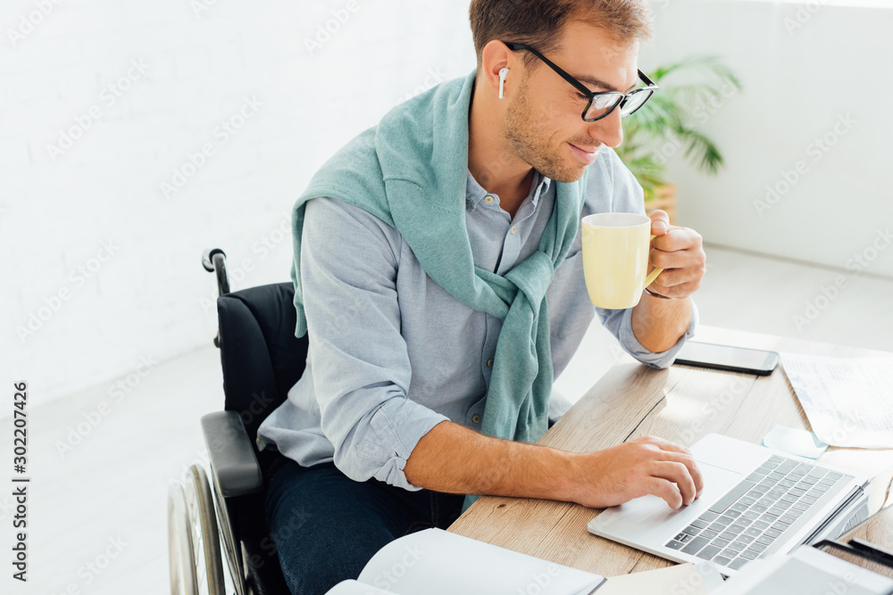 Casual businessman in wheelchair using wireless earphones and laptop while holding cup