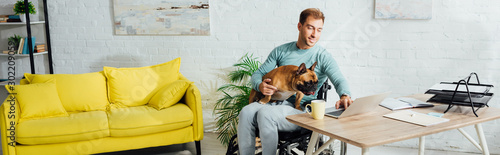 Disabled man holding french bulldog on knees and working on laptop, panoramic shot