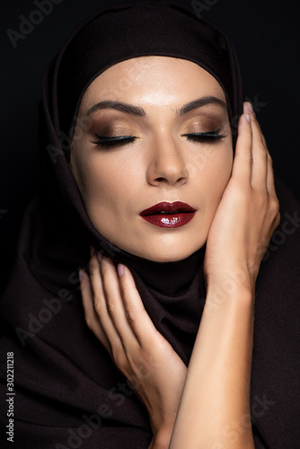 young Muslim woman in hijab with closed eyes, smoky eyes and red lips isolated on black
