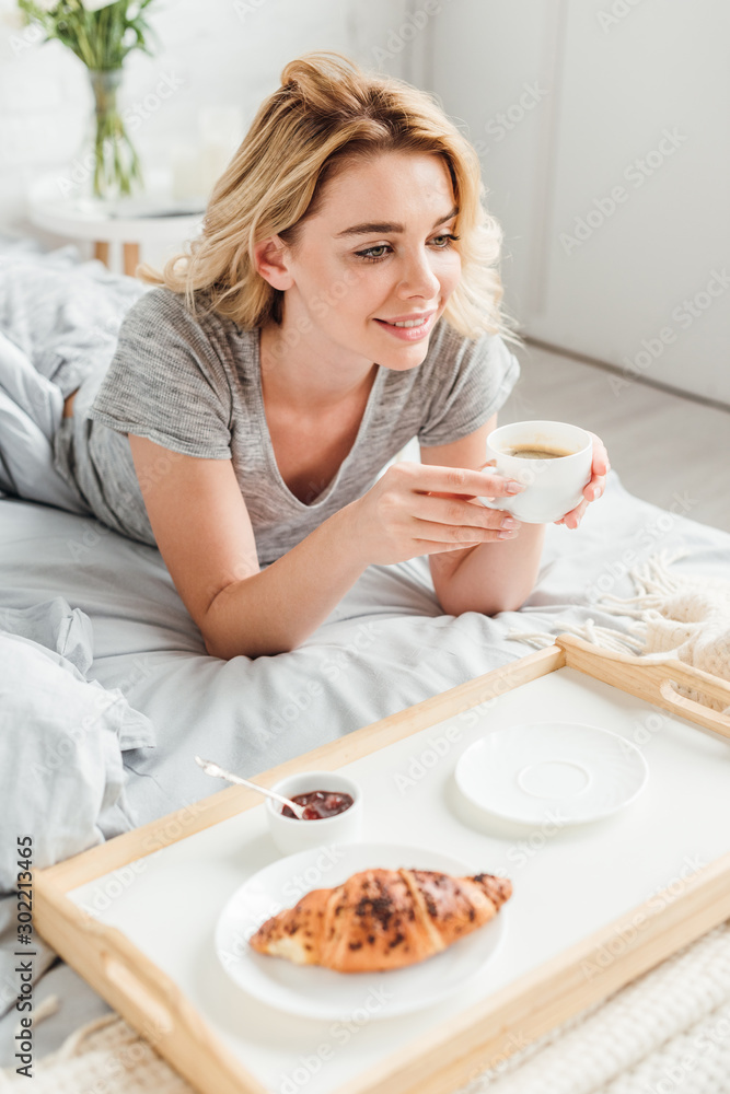 selective focus of cheerful girl holding cup of coffee near tray with tasty croissant on bed selective focus of cheerful girl holding cup of coffee near tray with tasty croissant on bed