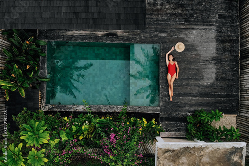 Aerial drone photo of happy Woman in red swimsuit relaxing near private pool with flowers and greenery around, Bali. Tropical background and travel concept.