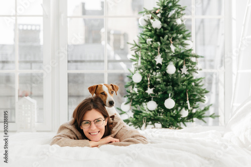 Pleased good looking young woman with dark hair wears round optical spectacles, smiles gently, lie on bed with domestic pet, have rest during winter holidays, green decorated New Year tree behind