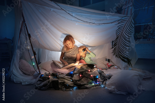 Mother and daughter sitting in a teepee, reading stories with the flashlight in dark room with toys and pillows. Caucasian models. Home comfort, family, love, Christmas holidays, storytelling time.