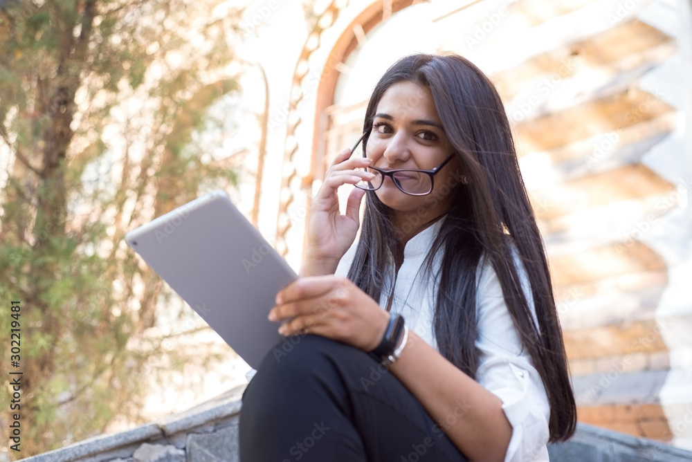 Fototapeta premium portrait of a beautiful indian girl. Business woman, wearing glasses, smiling, sitting on the steps of an office building, holding a tablet in her hand