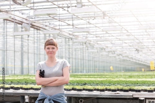 Portrait of confident young female botanist standing with arms crossed against herbs in greenhouse