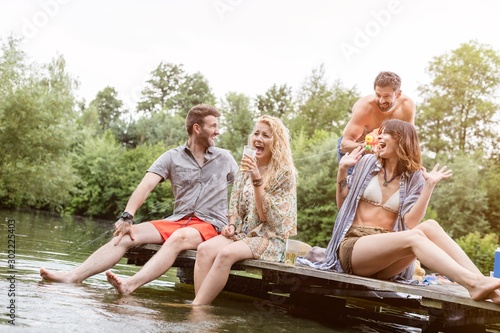 Cheerful shirtless man spraying water with squirt gun on friends sitting at pier over lake