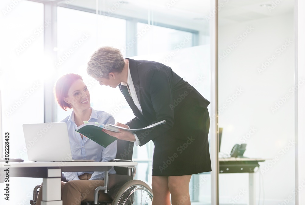 Mature businesswoman helping colleague sitting on wheel chair in office Mature businesswoman helping colleague sitting on wheel chair in office