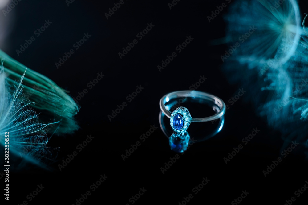 white gold ring with sapphire stone, against a black mirror background, in soft pastel blue tones. With a dandelion flower. Jewelry white gold ring with sapphire stone, against a black mirror background, in soft pastel blue tones. With a dandelion flower. Jewelry