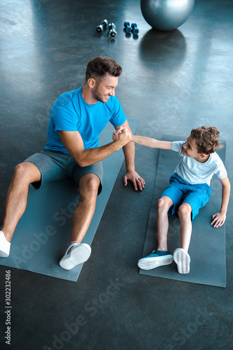 overhead view of happy father and son holding hands while sitting on fitness mats