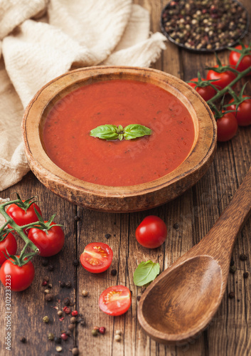 Wooden plate of creamy tomato soup with wooden spoon, pepper and kitchen cloth on wooden background with raw tomatoes.