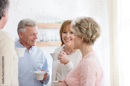 Happy mature friends holding drinks while talking in living room at home