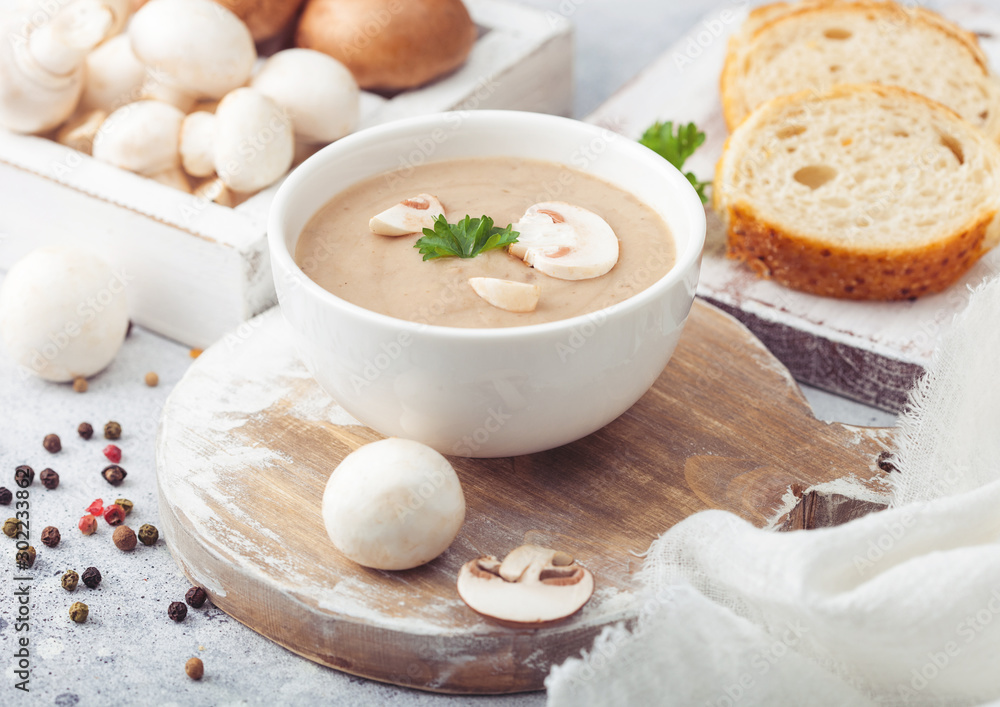 White bowl plate of creamy chestnut champignon mushroom soup on white kitchen background and box of raw mushrooms and fresh bread. White bowl plate of creamy chestnut champignon mushroom soup on white kitchen background and box of raw mushrooms and fresh bread.