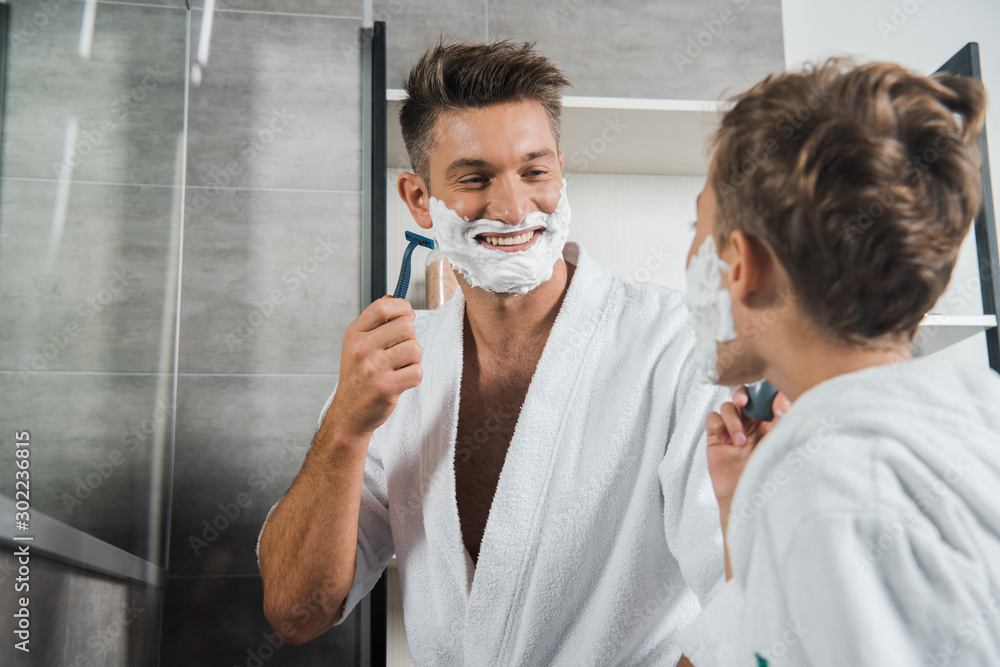 selective focus of happy father with shaving foam on face looking at son in bathroom