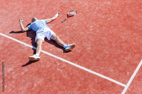 Full length of disappointed mature man lying by tennis racket on court during summer