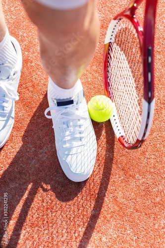 Low section of senior man standing with tennis racket and ball on red court © moodboard