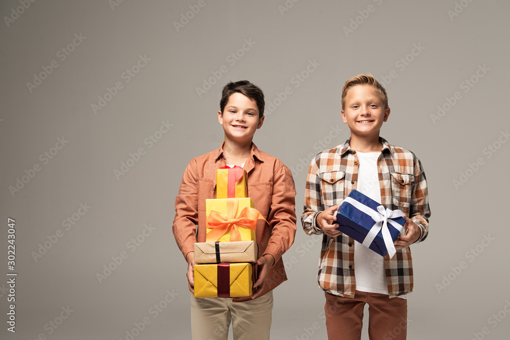 two happy brothers holding multicolored gift boxes and smiling at camera isolated on grey