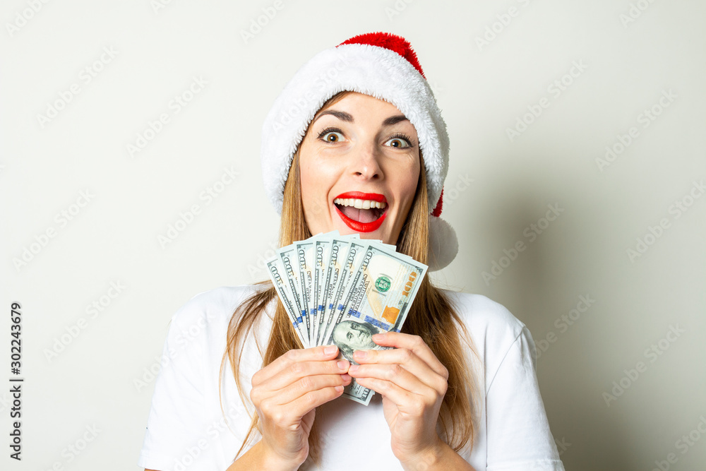 Young woman with a surprised face in a Santa Claus hat is holding a wad of money in his hands on a white background. Christmas sale concept, black friday, discount, rich, win, bet Young woman with a surprised face in a Santa Claus hat is holding a wad of money in his hands on a white background. Christmas sale concept, black friday, discount, rich, win, bet