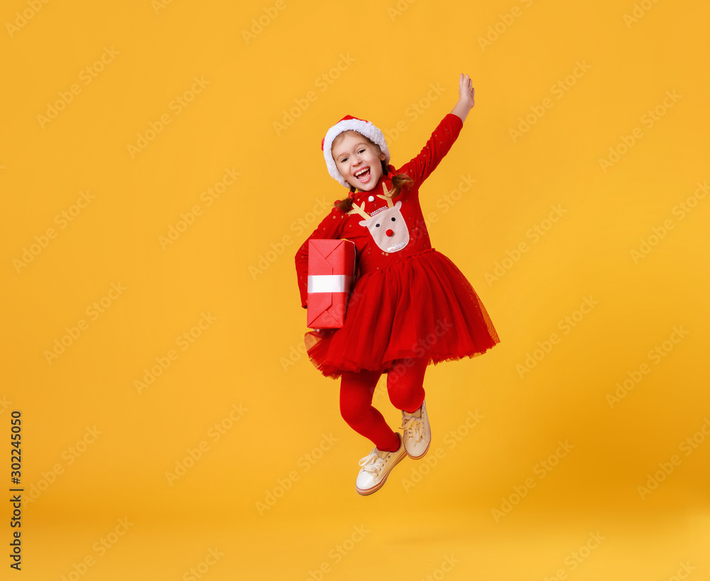 happy funny child girl in red Christmas reindeer costume jumping  with gift on yellow   background.