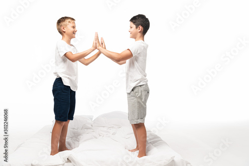 Fototapeta Naklejka Na Ścianę i Meble -  two smiling brothers clapping hands while standing on bed isolated on white