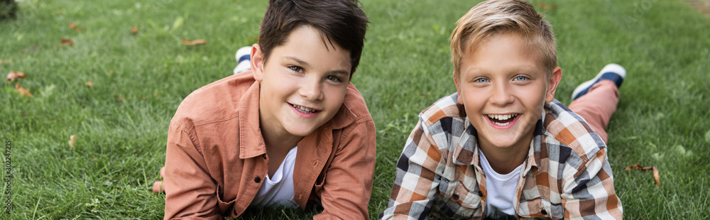 panoramic shot of two cheerful brothers lying on green grass and smiling at camera panoramic shot of two cheerful brothers lying on green grass and smiling at camera