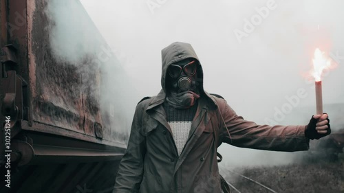 Hooded man in protective gas mask walks through abandoned carriages at destroyed train station holding burning red signal fire in hand. Apocalypse, survivor after nuclear or chemical disaster