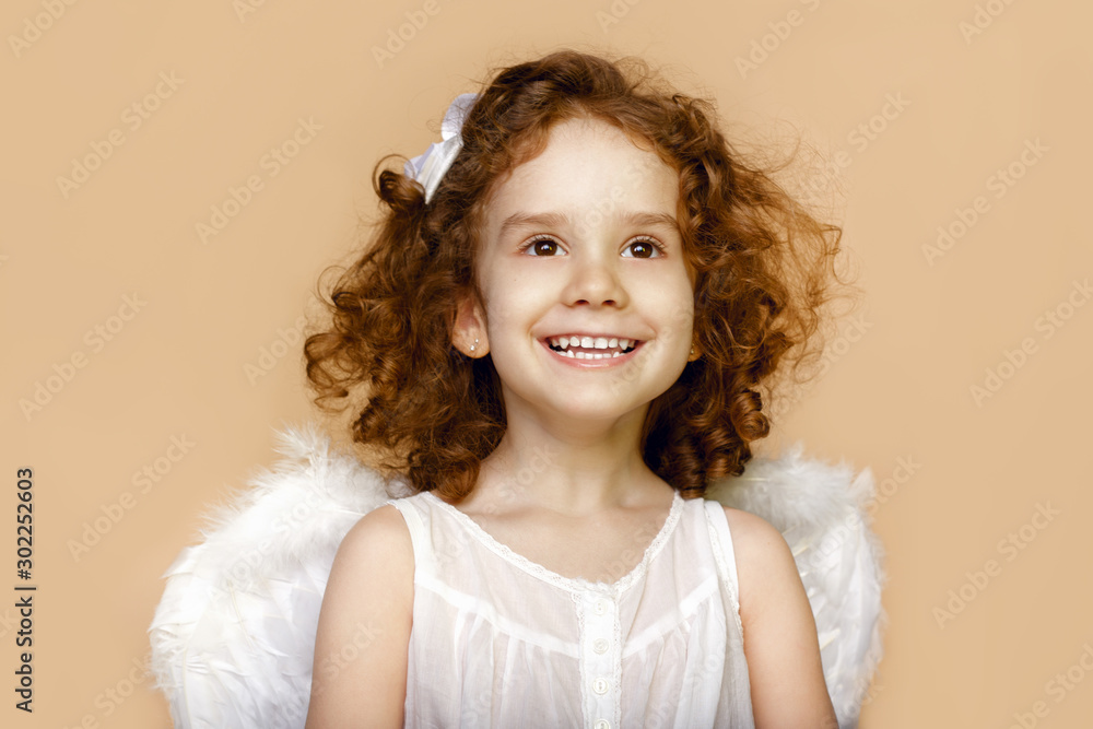 Little girl smiling, looking at a side, with wings in white dress, over beige background. Valentines day cupid. Little girl smiling, looking at a side, with wings in white dress, over beige background. Valentines day cupid.