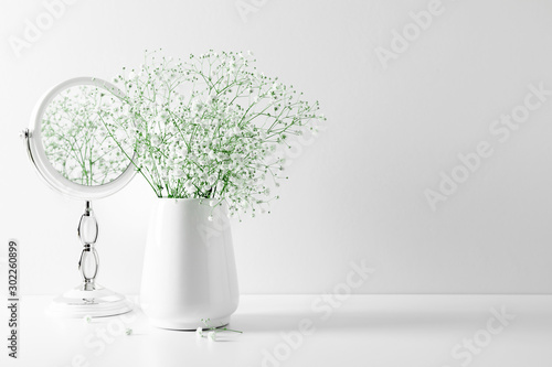 Elegant floral soft white composition, mirror, white flowers. Beautiful white gypsophila flower in vase on white wall background.