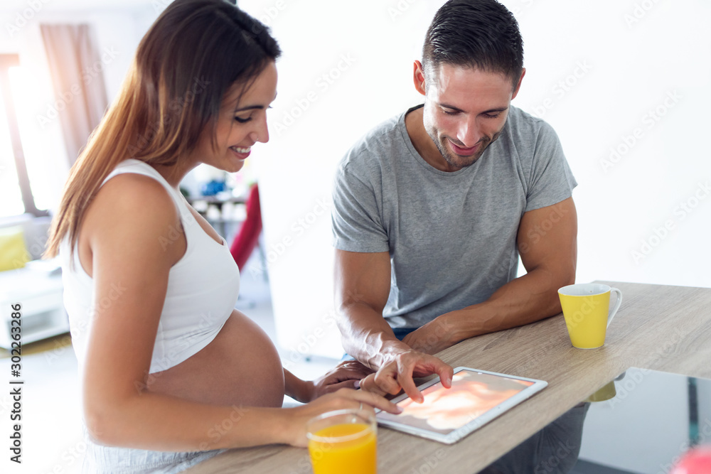 Pretty young pregnant couple looking at their baby's ultrasound on the digital tablet in the kitchen at home.
