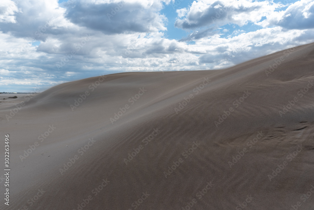 Sand forming dunes in the middle of the desert with a sky with clouds. Brown, blue and gray colors