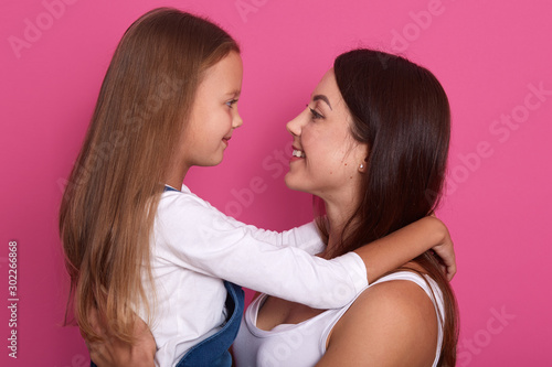 Excited mother and cute kid girl looking on each other with charming smiles, weaeing white shirts, having long hair, posing isolated over pink background, adorable female misses her liitle daughter.