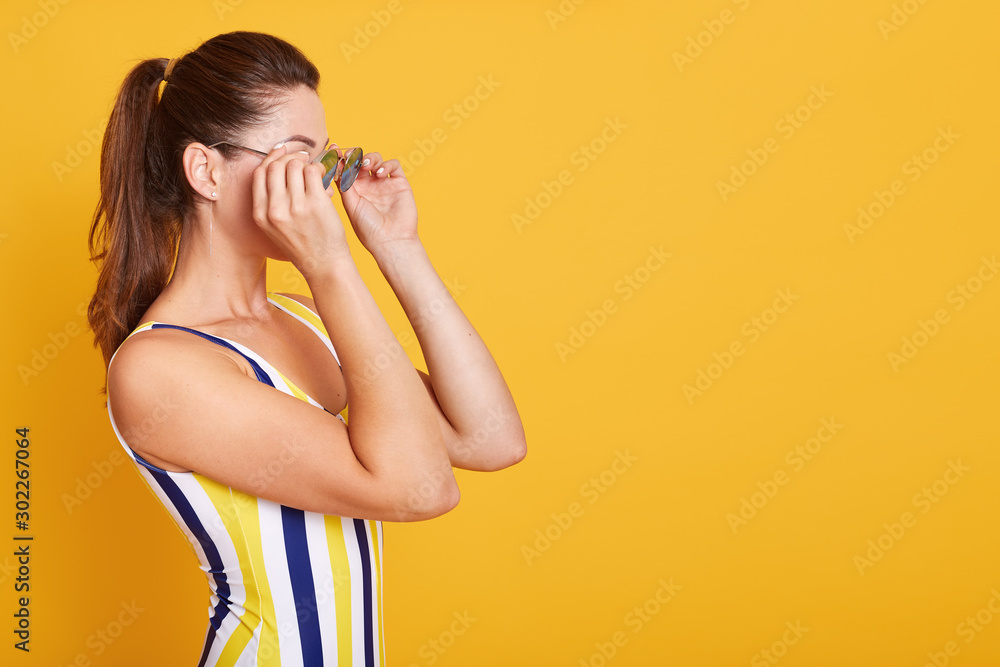 Close up portrait of young woman dressed swimsuit with stripes, holds sunglasses with both hands, posing isolated on yellow background, female with dark hair and ponitail. Copy space for advertisment. Close up portrait of young woman dressed swimsuit with stripes, holds sunglasses with both hands, posing isolated on yellow background, female with dark hair and ponitail. Copy space for advertisment.