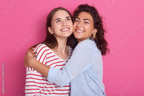 Two pretty sisters or friends women with dark hair, wearing casual shirts. Girls hugging over pink background in studio, ladies having happy expression, celebrating something, look with charming smile photo