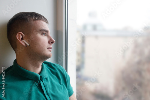 Sad pensive guy is listening to music in new wireless earphones or headphones, looking out window, sitting on a windowsill at home. Young man is dreaming, thinking about life. Sad mood. Heartbreak.