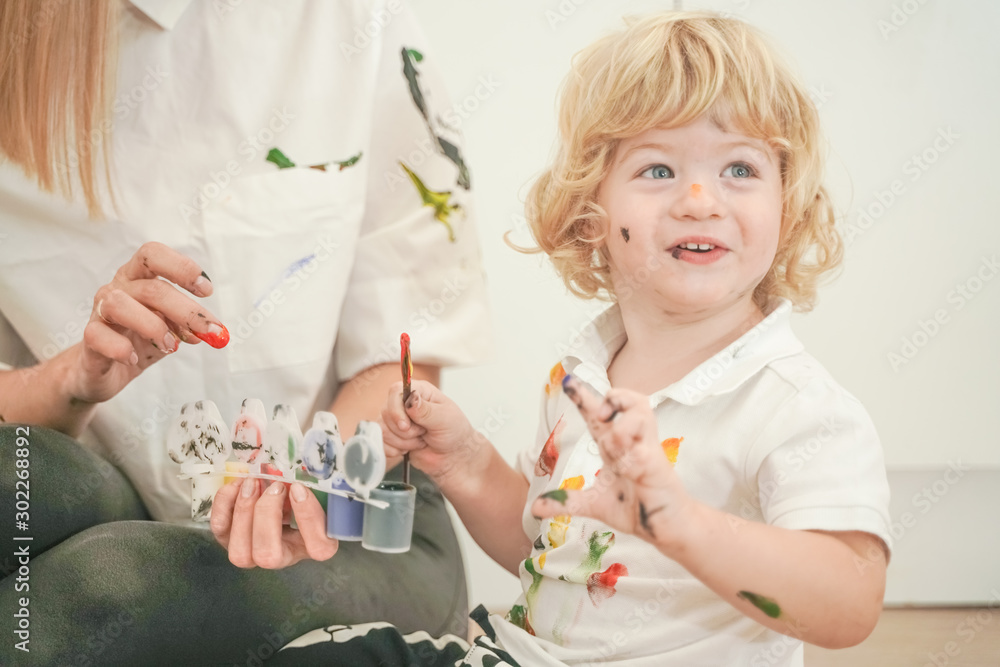 a cheerful mother and a joyful child sit on the floor in white t-shirts and paint each other with paints a cheerful mother and a joyful child sit on the floor in white t-shirts and paint each other with paints