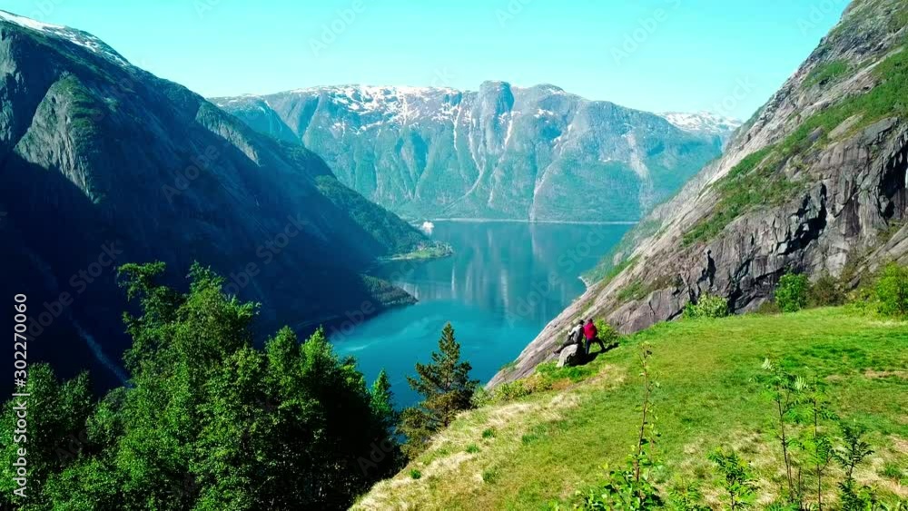 A drone areal capture of a fjord in Norway. Flight over a couple standing at the edge of the mountain. Long view on the fjord. Beautiful clarity in the sky. Lush green slopes, deep blue color of water