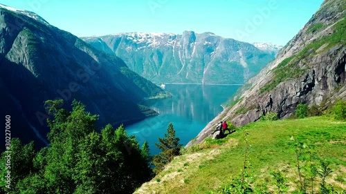 A drone areal capture of a fjord in Norway. Flight over a couple standing at the edge of the mountain. Long view on the fjord. Beautiful clarity in the sky. Lush green slopes, deep blue color of water