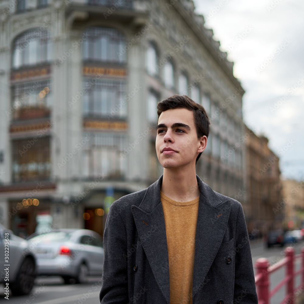 Obraz premium Handsome stylish fashionable man, brunette in elegant gray coat, stands on street in historical center of St. Petersburg. Young man with dark hair, thick eyebrows.