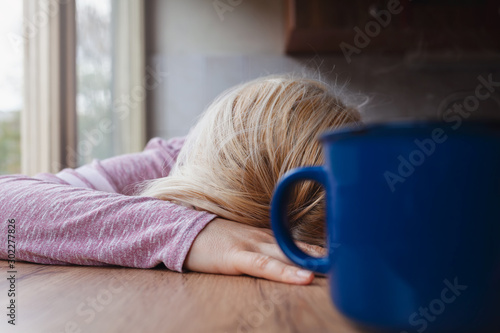 Blonde woman resting her head on a kitchen table, blue cup of coffee in front of her